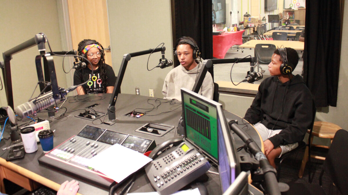 Three teens wearing headphones speaking into microphones in a radio studio