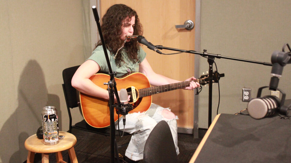 An adult playing acoustic guitar and a harmonica into a microphone in a radio studio