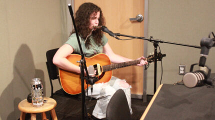 An adult playing acoustic guitar and a harmonica into a microphone in a radio studio