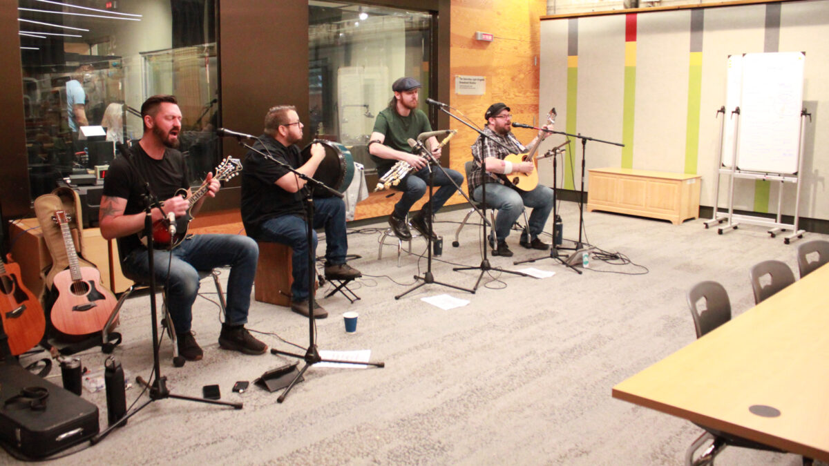 An adult playing a mandolin and singing into a microphone, an adult playing a bodhrán, an adult playing uilleann pipes, and an adult playing a bouzouki and singing into a microphone in front of a radio studio