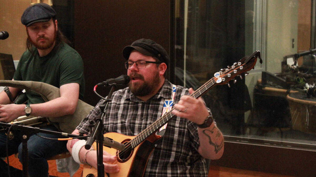 An adult playing uilleann pipes and an adult playing a bouzouki and singing into a microphone in front of a radio studio