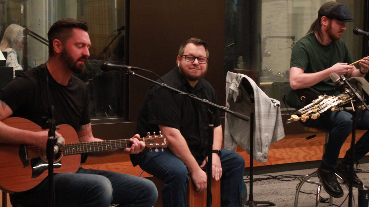An adult playing an acoustic guitar, an adult playing a cajon, and an adult playing a whistle in front of a radio studio