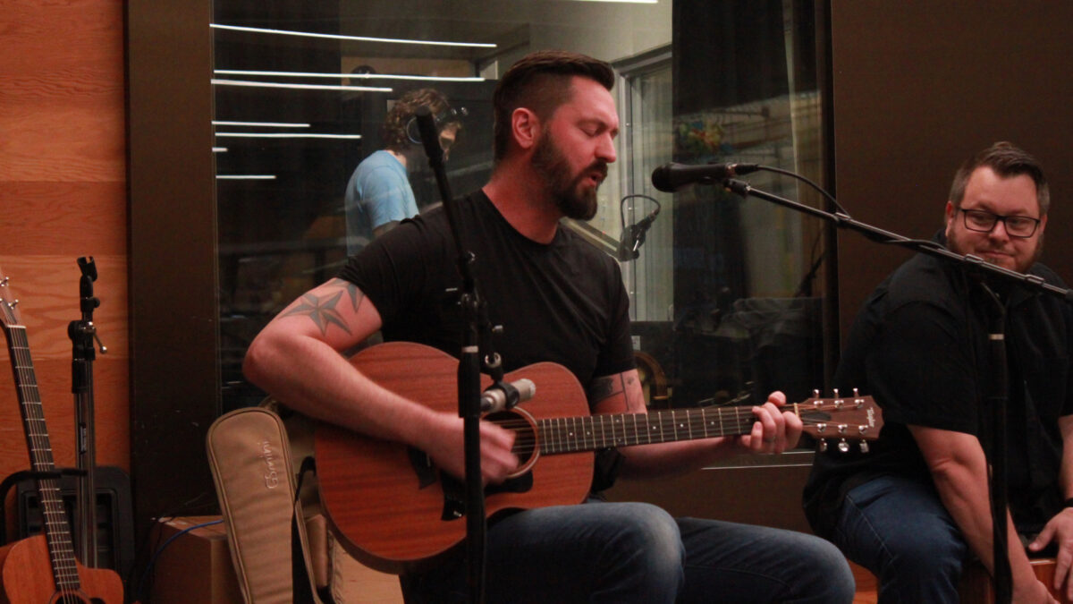 An adult playing an acoustic guitar and singing into a microphone and an adult playing a cajon in front of a radio studio