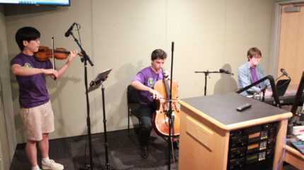 An adult playing a violin, an adult playing a cello, and an adult playing a piano keyboard in a radio studio
