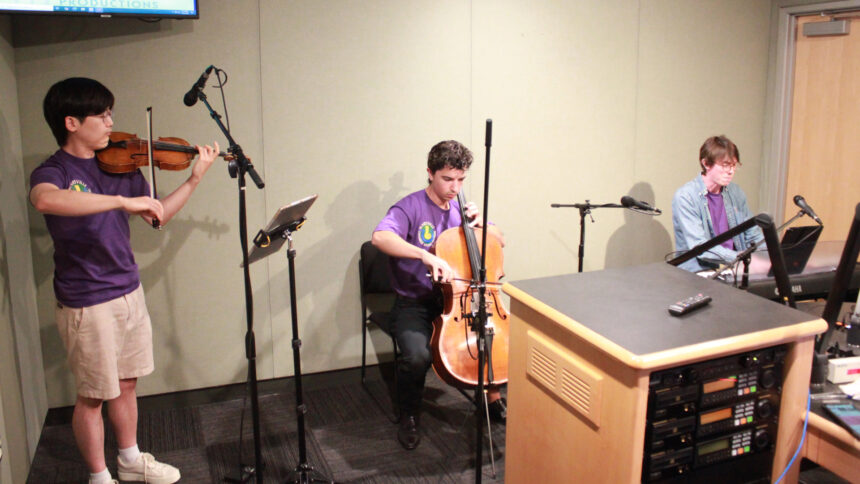 An adult playing a violin, an adult playing a cello, and an adult playing a piano keyboard in a radio studio