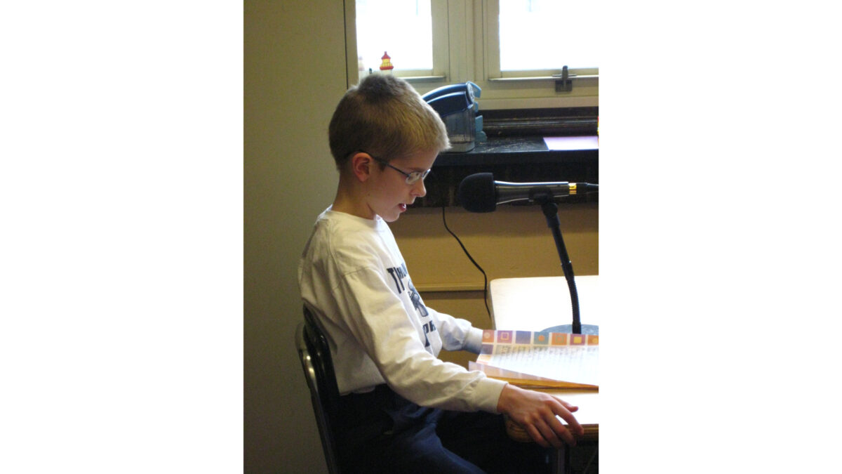 A youth reading into a microphone while sitting at a classroom desk
