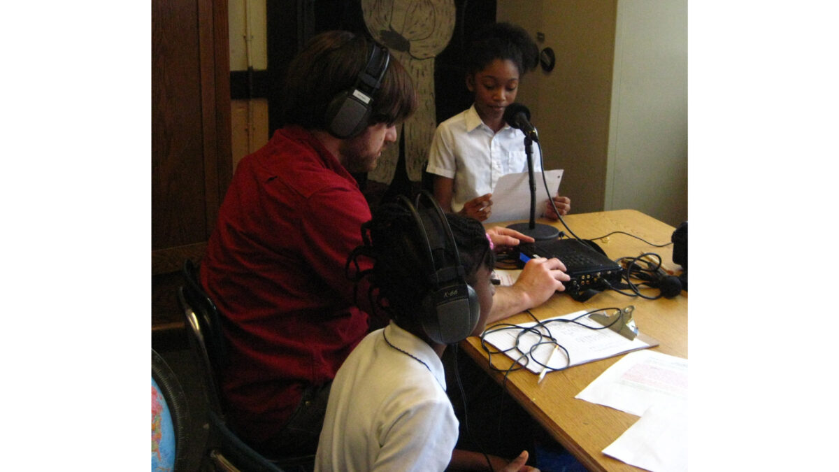 An adult and two youth wearing headphones and sitting at a classroom table, one youth sitting behind a microphone and holding a piece of paper
