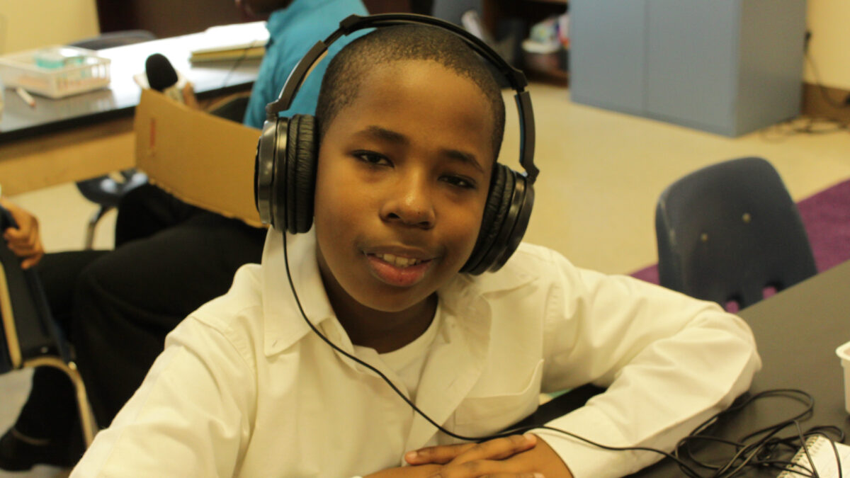 A youth wearing headphones smiling at the camera while sitting at a classroom table