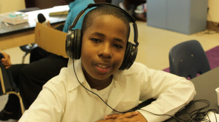 A youth wearing headphones smiling at the camera while sitting at a classroom table
