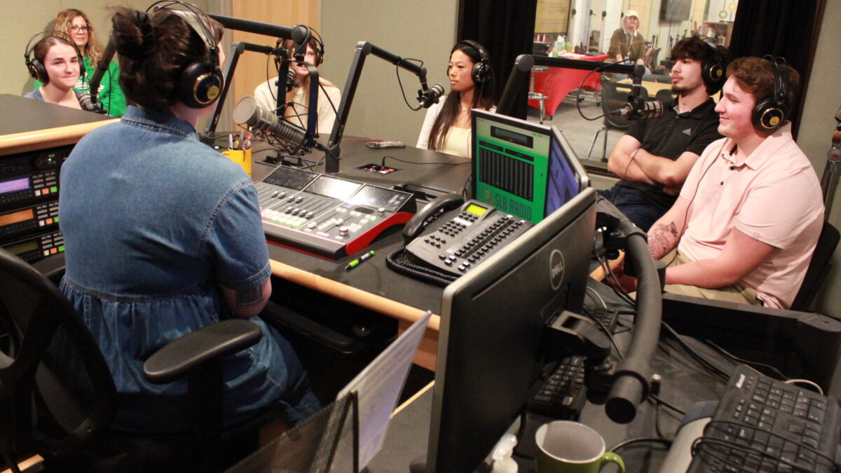 Five teens wearing headphones sitting behind microphones and an adult wearing headphones sitting behind a microphone and an audio mixer in a radio studio