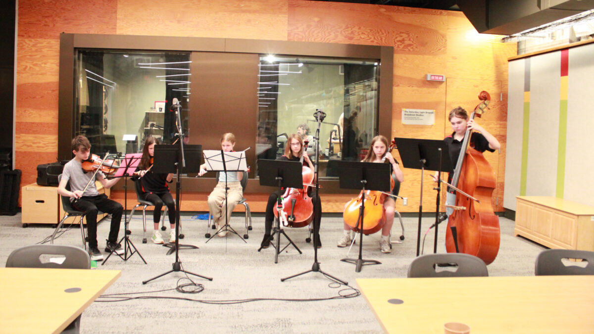 Three youth playing violins, two youth playing cellos, and a youth playing string bass, all looking at sheet music on music stands in front of a radio studio
