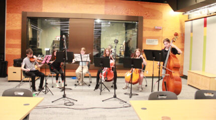 Three youth playing violins, two youth playing cellos, and a youth playing string bass, all looking at sheet music on music stands in front of a radio studio