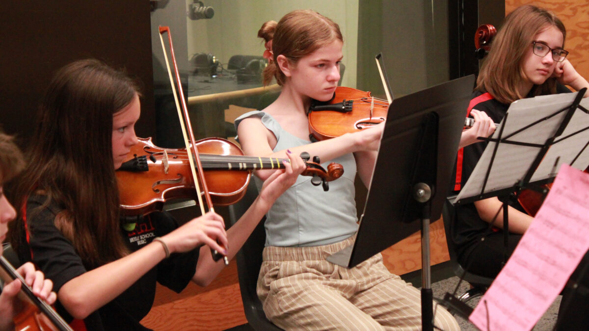 Two youth playing violin and a youth holding a cello, all looking at sheet music on music stands