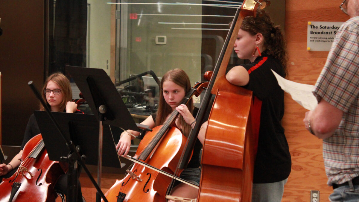 A youth holding a cello, a youth playing a cello while looking at sheet music on a music stand, and a youth holding a string bass