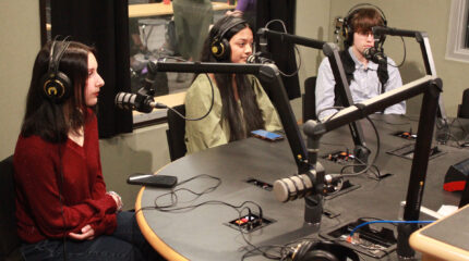 Three teens wearing headphones sitting behind microphones in a radio studio