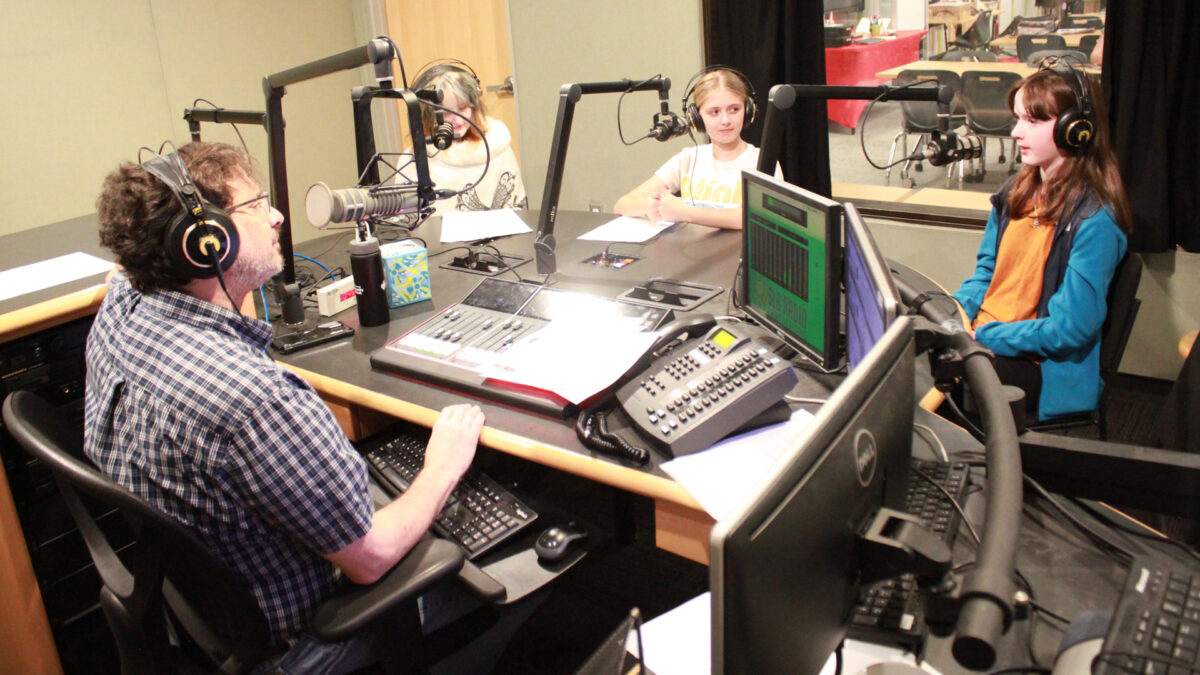 Three teens wearing headphones speaking into microphones being interviewed by an adult wearing headphones sitting behind a microphone and an audio mixer in a radio studio