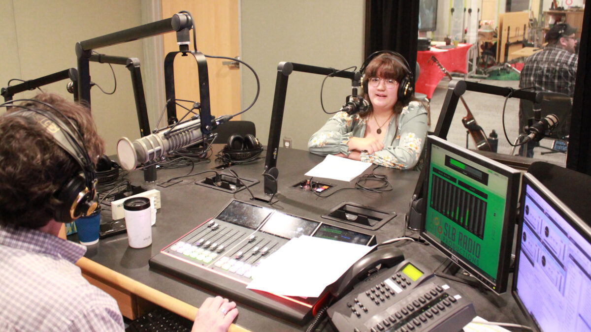 A teen wearing headphones speaking into a microphone being interviewed by an adult wearing headphones sitting behind a microphone and an audio mixer in a radio studio