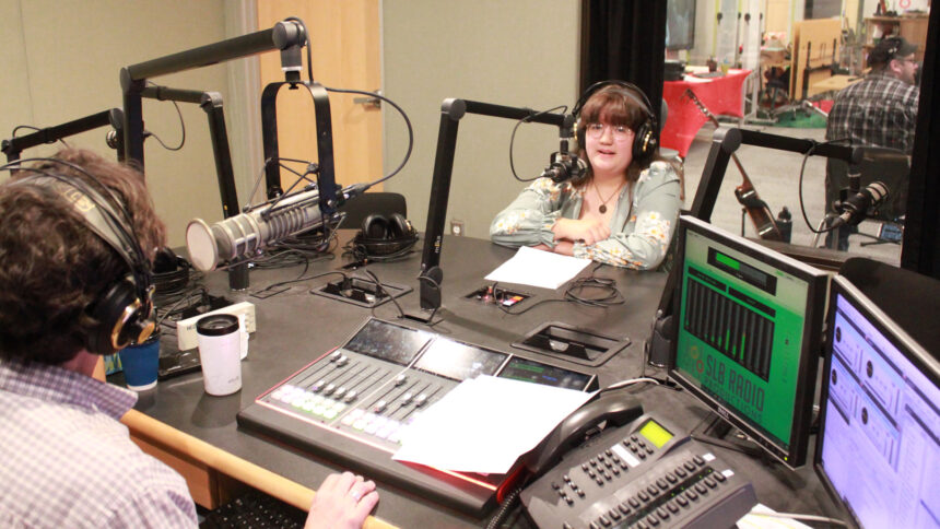 A teen wearing headphones speaking into a microphone being interviewed by an adult wearing headphones sitting behind a microphone and an audio mixer in a radio studio