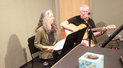 An adult playing a drum and an adult playing acoustic guitar and singing into a microphone in a radio studio