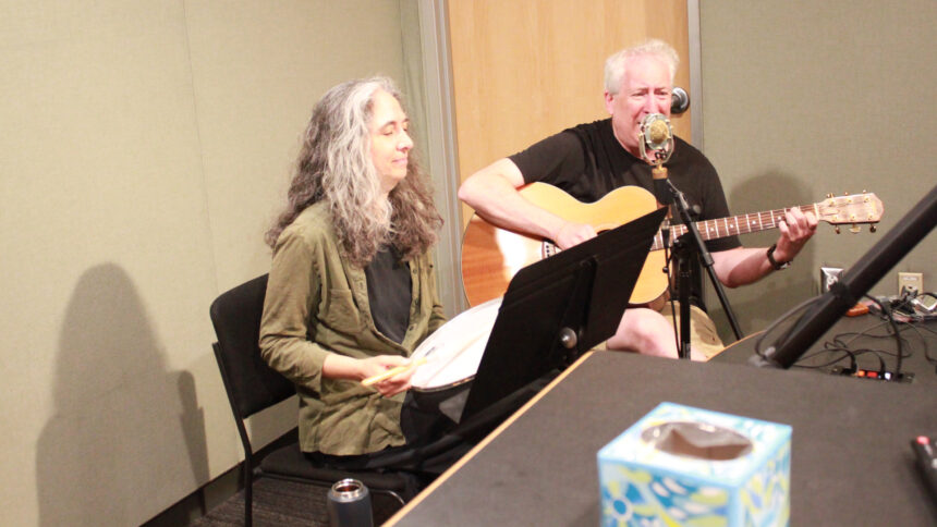 An adult playing a drum and an adult playing acoustic guitar and singing into a microphone in a radio studio