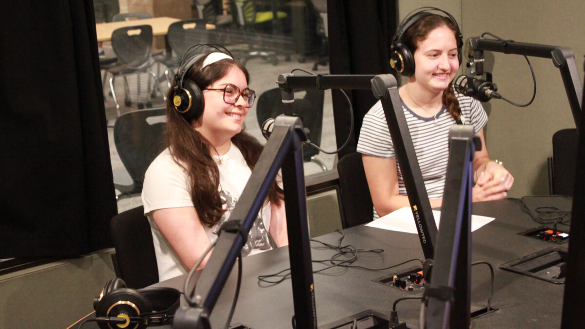 Two teens wearing headphones smiling while sitting behind microphones in a radio studio