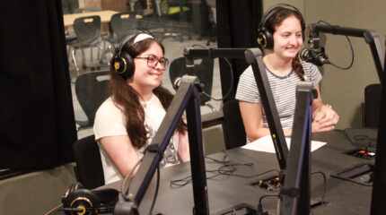 Two teens wearing headphones smiling while sitting behind microphones in a radio studio