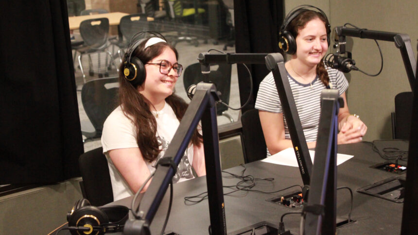 Two teens wearing headphones smiling while sitting behind microphones in a radio studio