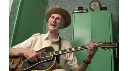 An adult smiling at the camera and playing an acoustic guitar in a green room
