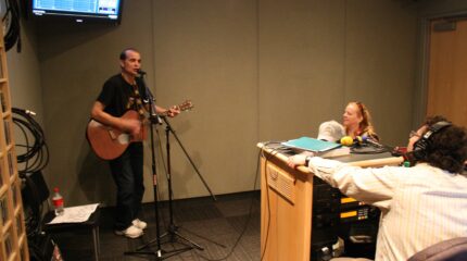 An adult playing an acoustic guitar and singing into a microphone as two adults listen in a radio studio