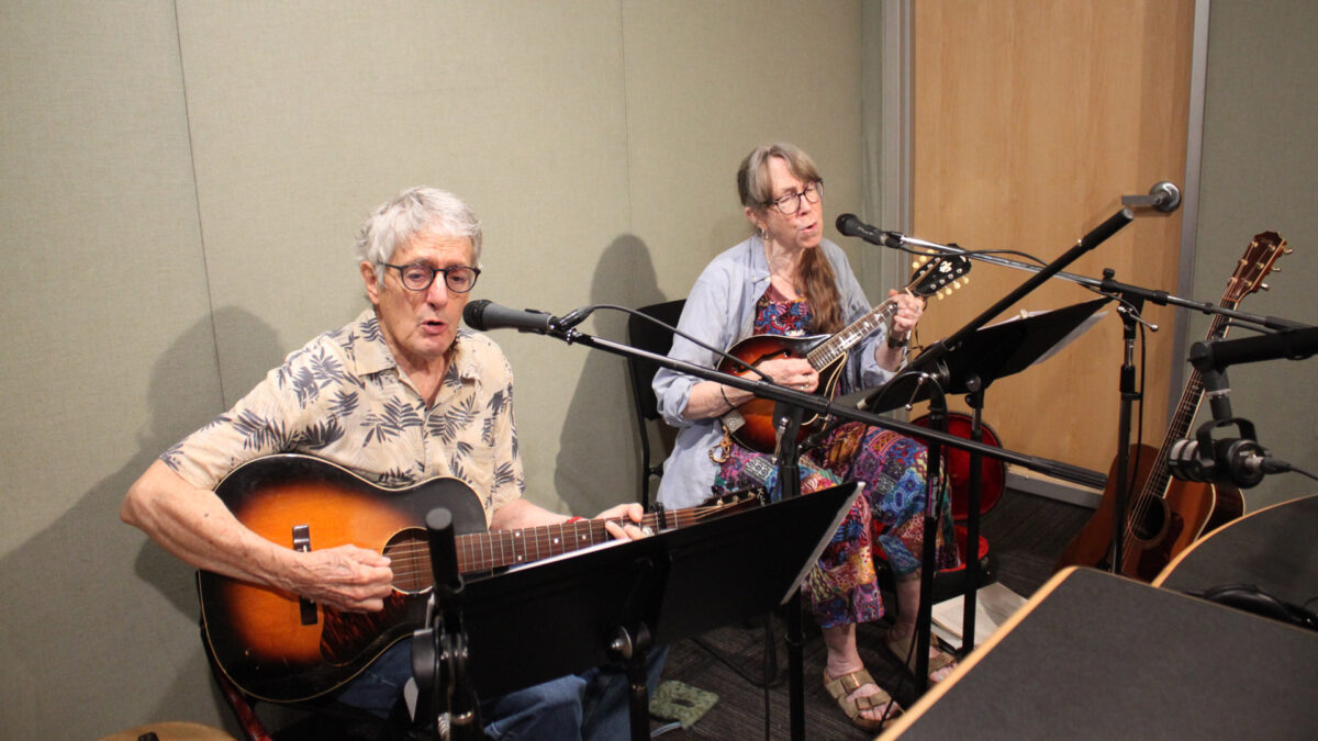 Two adults singing into microphones in a radio studio, one playing acoustic guitar, one playing mandolin