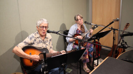 Two adults singing into microphones in a radio studio, one playing acoustic guitar, one playing mandolin