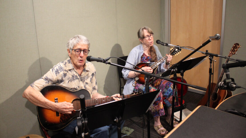 Two adults singing into microphones in a radio studio, one playing acoustic guitar, one playing mandolin