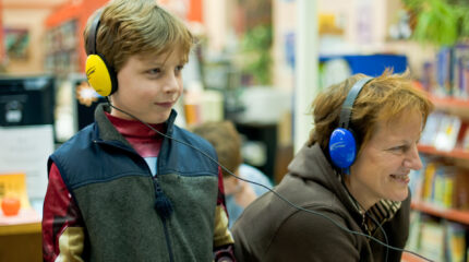 A youth and an adult wearing headphones and smiling in a library
