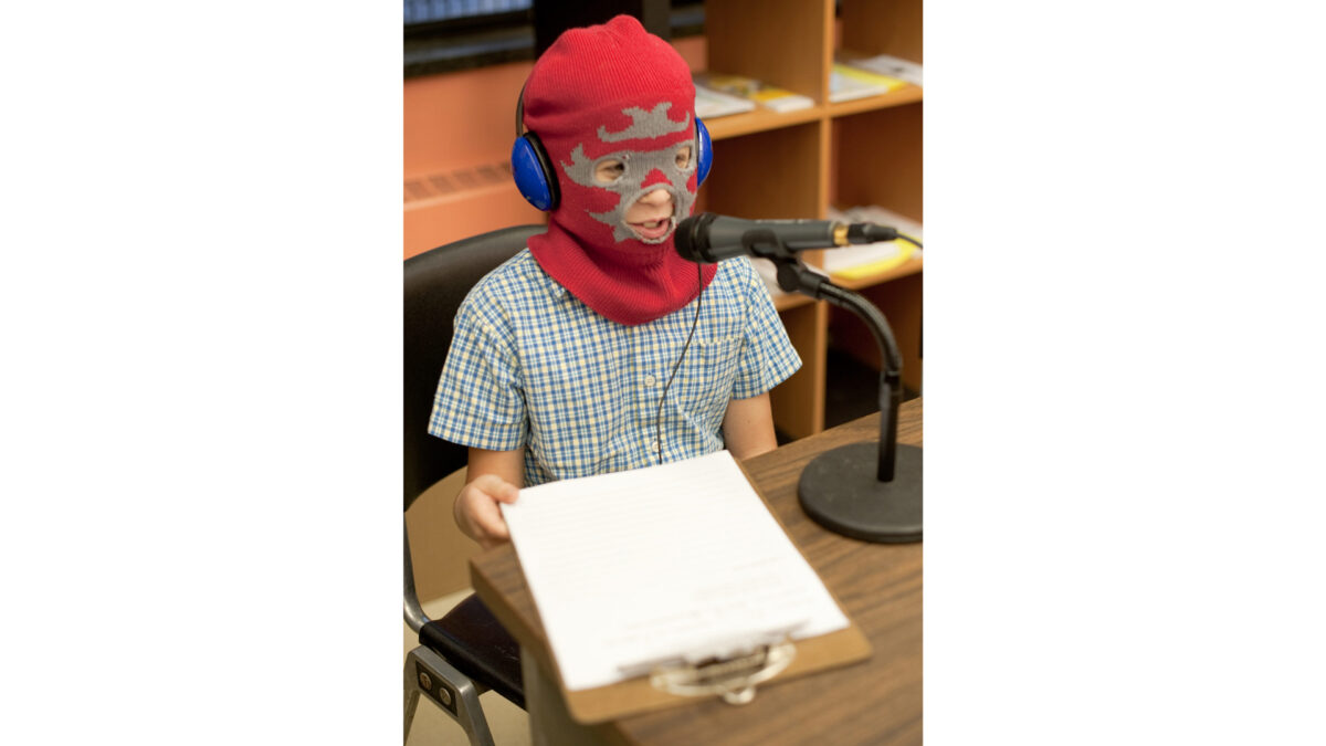 A youth wearing a ski mask and headphones speaking into a microphone and holding a clipboard