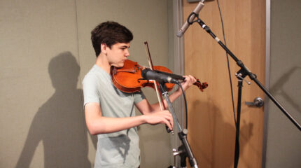 A teen playing a viola standing behind microphones in a radio studio