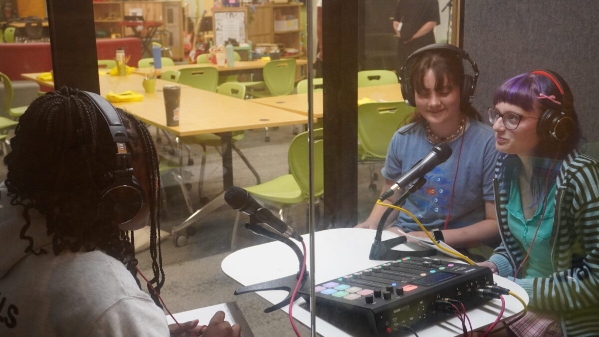 Three teens in an audio recording booth recording a conversation together.
