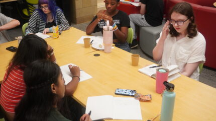 A group of teens at a table writing in notebooks and thinking intently.