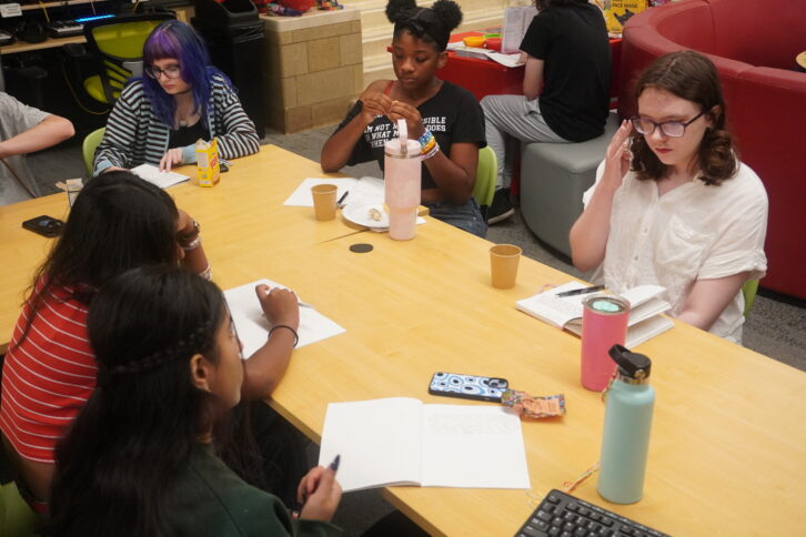A group of teens at a table writing in notebooks and thinking intently.