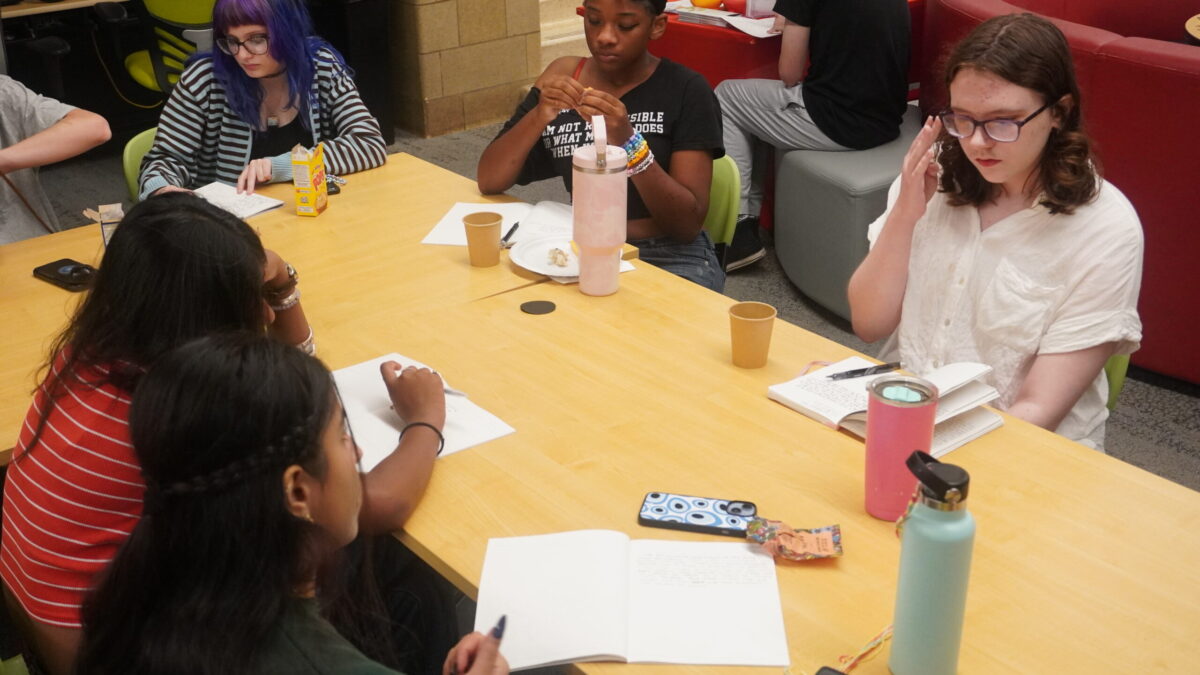 A group of teens at a table writing in notebooks and thinking intently.