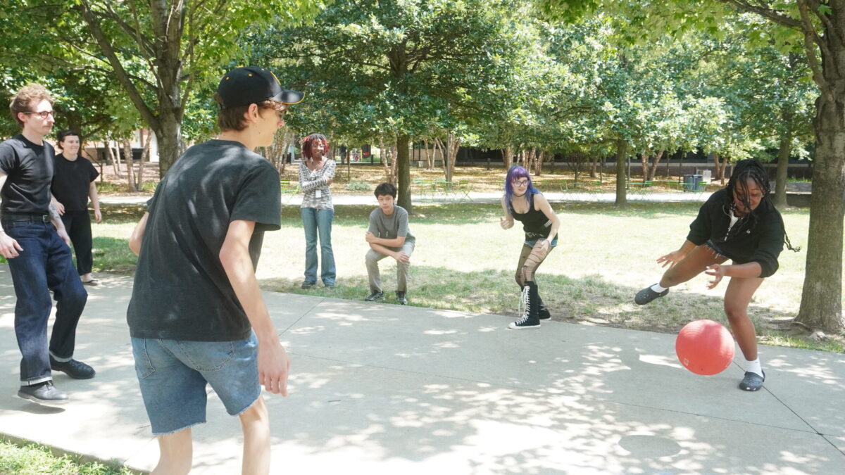 A group of teens and instructors outdoors as an inflated ball bounces across a sidewalk. A teen reaches to relay the ball.