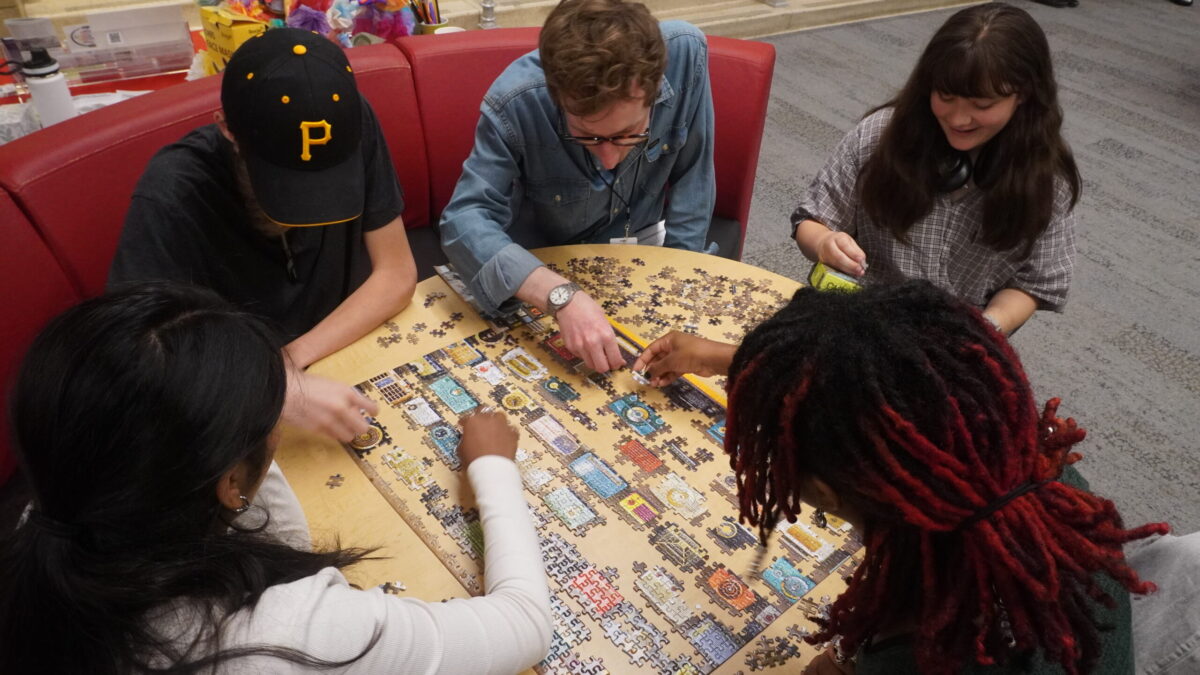 A group of teens and adults assemble pieces of a jigsaw puzzle at a table.