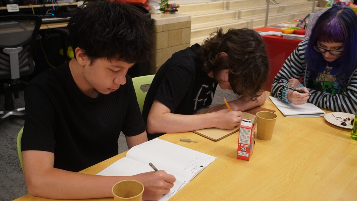 Three teens at a table writing in notebooks
