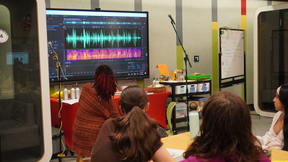 A group of teens and their instructor look upon an audio editing software on a large screen TV.