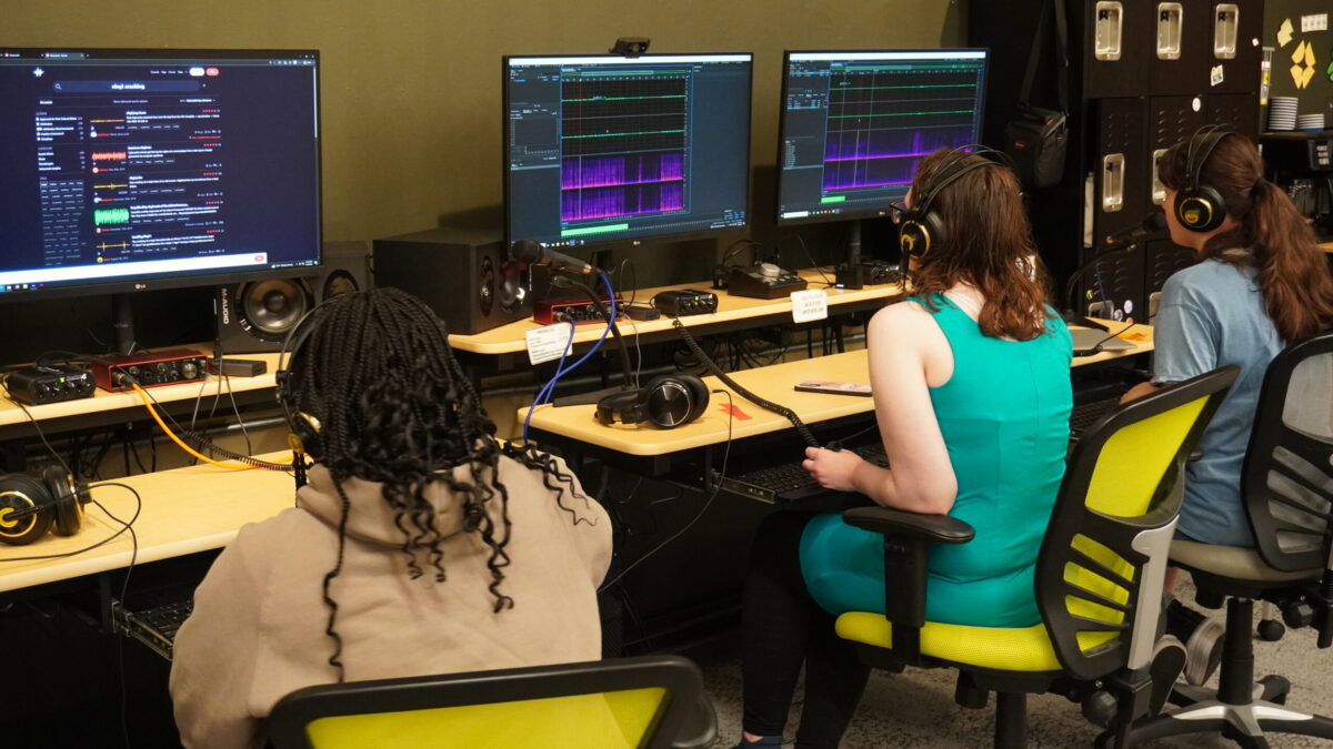 Three teens sitting at computers, wearing headphones, and editing audio.