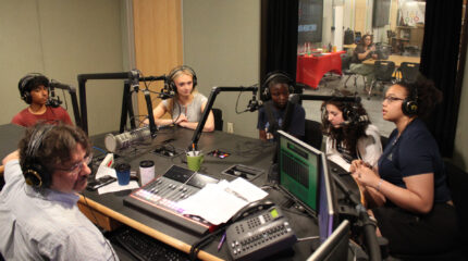 Five teens wearing headphones speaking into microphones being interviewed by an adult wearing headphones sitting behind an audio mixer in a radio studio