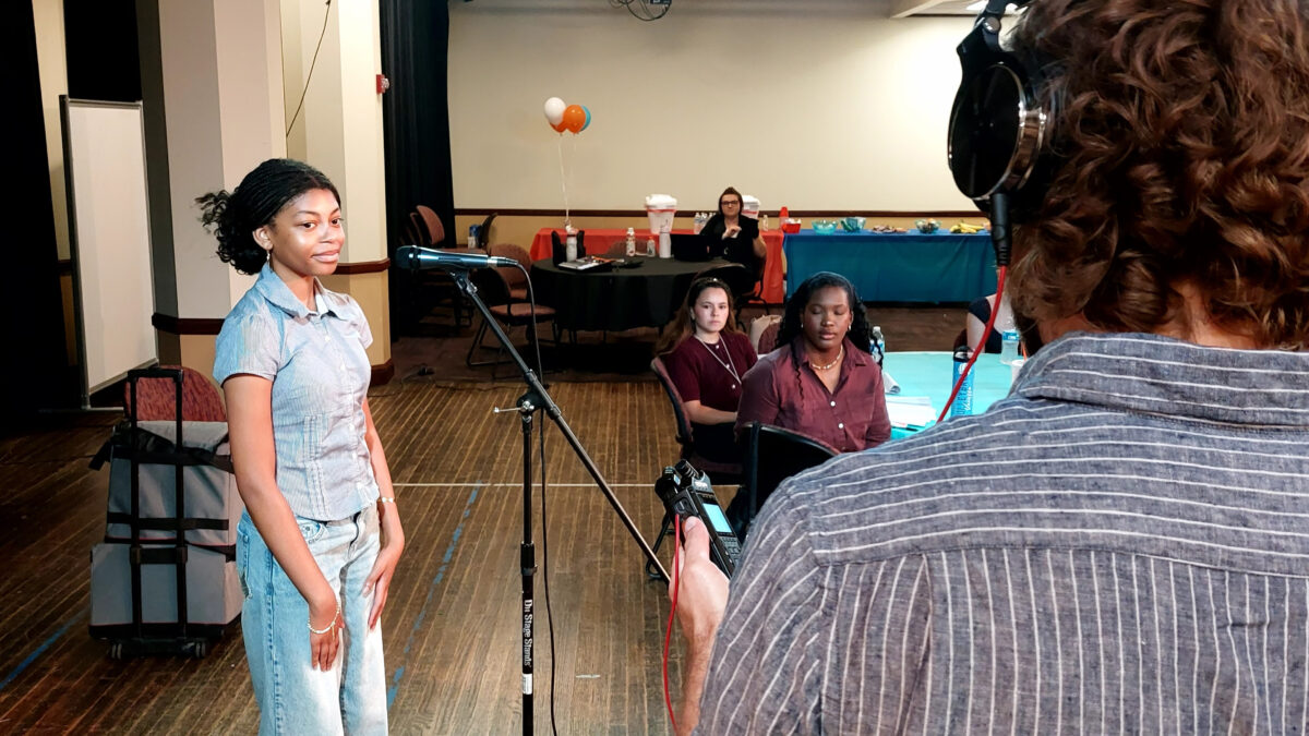 A teen standing at a microphone watching an adult wearing headphones operate an audio recorder while other teens look on.