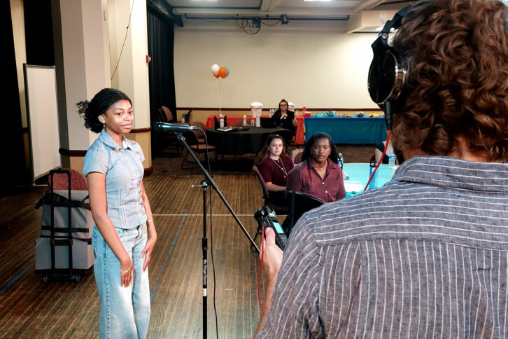 A teen standing at a microphone watching an adult wearing headphones operate an audio recorder while other teens look on.