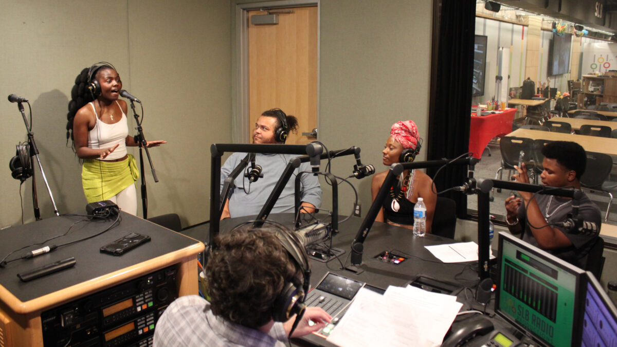 Once-On-This-Island-1 An adult wearing headphones singing into a microphone as four adults wearing headphones listen in a radio studio