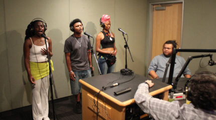 Three adults wearing headphones singing into microphones as two adults wearing headphones listen in a radio studio