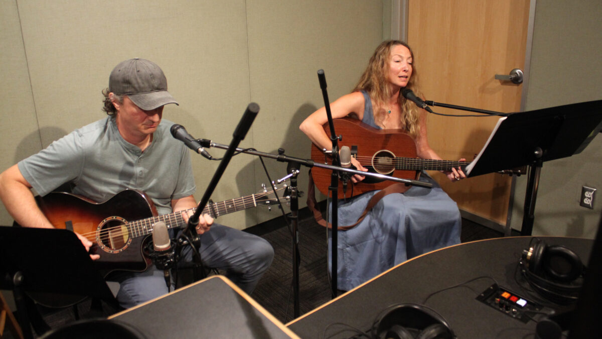 Two adults playing acoustic guitar in a radio studio, one singing into a microphone